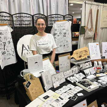 Allara Creative stall holder standing behind a market stall displaying illustrated tea towels, cards and prints.